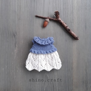 Op de afbeelding: Een gebreide poppenjurk met een blauwe bovenkant en een witte kanten rok. De jurk ligt op een grijze houten achtergrond met een klein takje en een eikel.
