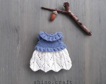 May include: A knitted doll dress with a blue bodice and a white lace skirt. The dress is on a gray wooden background with a small branch and acorn.