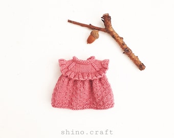 May include: A pink knitted dress with a ruffled collar. The dress is laying on a white surface with a small branch and acorn.