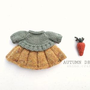 Puede incluir: Un vestido de punto para muñeca con un corpiño verde y una falda amarilla con motas de color. El vestido está sobre una superficie blanca junto a una pequeña zanahoria de punto.
