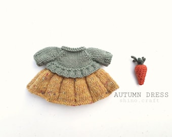 May include: A knitted doll dress with a green bodice and a yellow skirt with flecks of color. The dress is laying on a white surface next to a small knitted carrot.