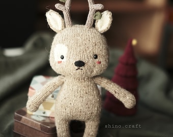 May include: A knitted deer toy with brown fur, white ears, and green antlers. The deer is standing on a green fabric surface with a small box and a red Christmas tree in the background.