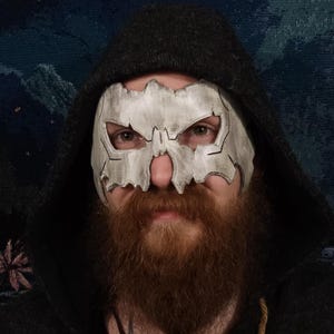 Może przedstawiać: Osoba w czarnym swetrze z kapturem i białą maską czaszki. Maska ma szczegółowy wzór z ostrymi krawędziami i fakturowaną powierzchnią.