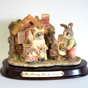 Puede incluir: Figura decorativa que representa una familia de conejos frente a una pequeña casa. Los conejos visten ropa e interactúan entre ellos. La figura está sobre una base de madera oscura con el texto "The Morning Post by Leonardo".