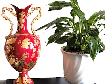 May include: A vibrant red ceramic vase with ornate gold handles and gold floral patterns, featuring a flared rim and a square base. Beside it sits a green leafy plant in a white, spiraled, footed ceramic pot, both on a black surface.