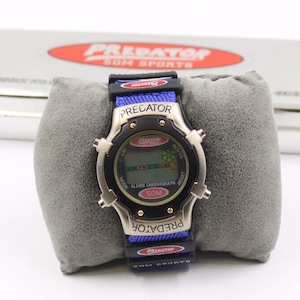 May include: A silver and black digital watch with a blue and black band, displayed on a gray cushion. The watch face displays the text "PREDATOR" and "50M SPORTS". A silver tin with the same logo is in the background.