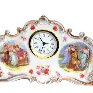 Puede incluir: Un reloj de sobremesa de porcelana blanca con bordes dorados, que presenta una esfera de cuarzo central con números romanos. El reloj está decorado con motivos florales coloridos y dos escenas pintadas ovaladas con grupos de figuras clásicas.