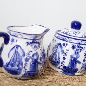 以下が含まれることがあります： 白と青のセラミック製クリーマーと砂糖入れのセット。風車、帆船、そして青色の「Holland」の文字が特徴的なオランダのデザインです。クリーマーは、湾曲したハンドルと注ぎ口が付いています。
