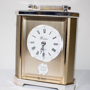 Può includere: Un orologio da tavolo dorato con un quadrante bianco e indici orari romani neri. L'orologio ha una cornice argentata e una maniglia argentata sulla parte superiore. Il quadrante dell'orologio riporta "Westclox" e "Tempus Fugit".