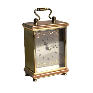 May include: A rectangular brass-coloured clock with a handle on top. The clock face is visible through a glass panel, displaying Roman numerals and clock hands. The clock has a decorative design around the face and a brown base.
