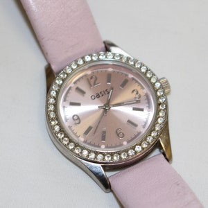 Puede incluir: Un reloj plateado con una correa de cuero rosa y un bisel engastado con cristales. La esfera del reloj es rosa con manecillas plateadas y números romanos. La palabra "oasis" está impresa en la esfera del reloj.
