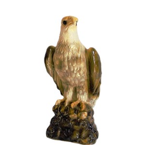 Könnte beinhalten: Eine Adlerfigur aus Keramik, die auf einem strukturierten, dunkelbraunen Felsensockel steht. Der Adler hat einen cremefarbenen Kopf und Brustbereich mit detaillierter Federstruktur, während die Flügel in glänzendem Olivgrün und Braun gehalten sind.