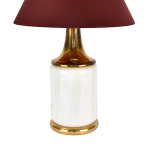 Puede incluir: Una lámpara de mesa de cerámica blanca con base dorada y pantalla de color burdeos.