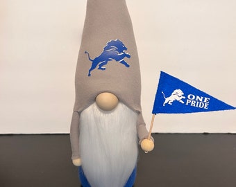 May include: A handmade gnome doll with a gray pointed hat featuring a blue lion logo. The gnome has a long white beard, a wooden nose, and a blue body. It holds a blue flag with a lion logo and the words "ONE PRIDE".