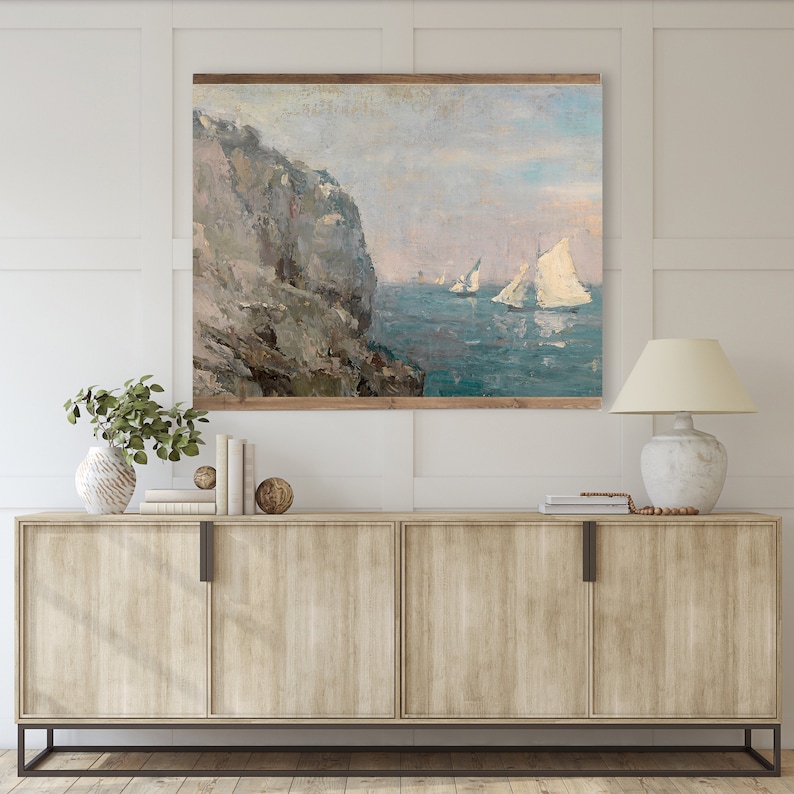 May include: A painting depicting a rocky cliff with sailboats in the distance. The sky is a light blue and the water is a turquoise colour. The painting is framed in a wooden frame.