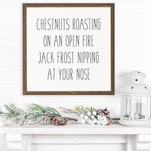 May include: A framed sign with the text "Chestnuts roasting on an open fire Jack Frost nipping at your nose" on a whitewashed wood background. A white lantern with black stars and a sprig of pine with white berries are on a white shelf below the sign.