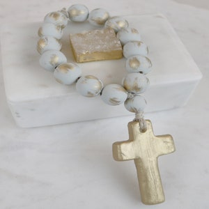 May include: A white and gold beaded rosary with a gold cross charm. The rosary is on a white marble surface.