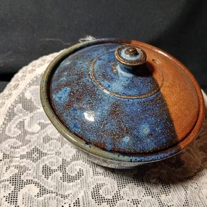 Può includere: Una ciotola rotonda in ceramica con coperchio abbinato, caratterizzata da una smaltatura bicolore. Un lato del coperchio e della ciotola è di un blu profondo e maculato, mentre l'altro lato è di un marrone terracotta liscio e uniforme. Un piccolo pomello si trova sul coperchio.