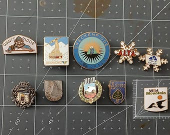 May include: A collection of enamel pins featuring various designs. Includes pins with text like "GIANT'S RIDGE", "Snowbird", "Breckenridge Colorado", "ALTA", "Welch Village", "Innsbruck", "SSM", "Aspen", and "Wild Mountain".