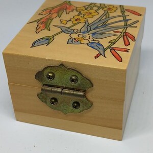 May include: A small wooden box with a hinged lid and a brass latch. The box is decorated with a painted floral design featuring blue, yellow, red, and green flowers.
