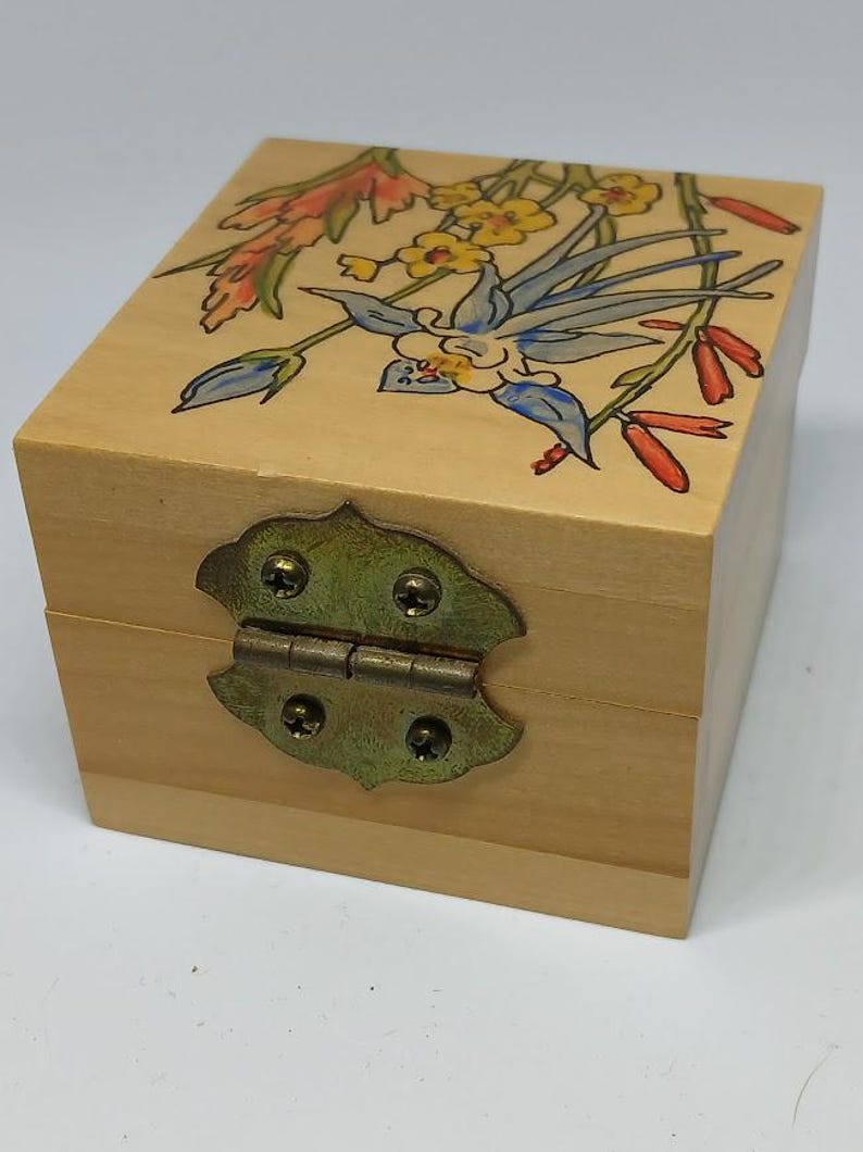 May include: A small wooden box with a hinged lid and a brass latch. The box is decorated with a painted floral design featuring blue, yellow, red, and green flowers.