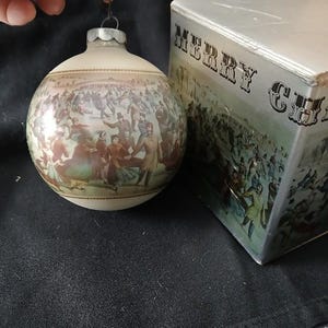 May include: Vintage Christmas ornament with a detailed illustration of people in period clothing. The ornament is round with a silver cap and a wire for hanging. A box with "MERRY CH" is next to the ornament.