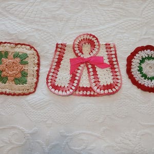 May include: Three crocheted items: a square coaster with a peach flower and green leaves, a white and red crocheted garment-shaped piece with a pink ribbon bow, and a round white coaster with a green ring and a red scalloped border edge.