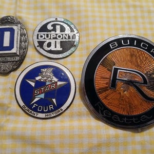 Può includere: Una collezione di emblemi di auto d'epoca, tra cui un emblema Durant blu e bianco, un emblema Dupont, un emblema Star Four Motors e un emblema Buick Reatta con dettagli arancioni e neri.