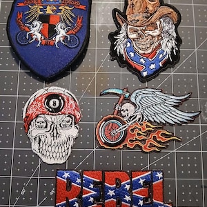 May include: A collection of embroidered patches featuring biker and rebel themes. Designs include a shield with text, a skull wearing a cowboy hat, a skull with a bandana, a skull riding a motorcycle, and the word "REBEL" in a star-spangled font.