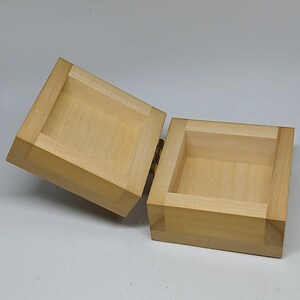 May include: A small, wooden box with a hinged lid. The box is made of light-colored wood and has a natural finish. The lid is open, revealing the empty interior of the box.