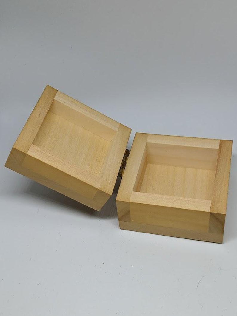 May include: A small, wooden box with a hinged lid. The box is made of light-colored wood and has a natural finish. The lid is open, revealing the empty interior of the box.
