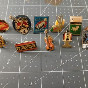 May include: A collection of vintage enamel pins featuring various designs. Includes pins with text like "VIET NAM 1975", "HELMET LAWS", "CLUB RACING", and "MAZDA". Other pins depict a rabbit, a violin, and a horse.