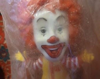 May include: A vintage Ronald McDonald figurine, encased in plastic. The clown figure has a white face, red nose and mouth, and orange hair. He wears a yellow vest, red and white striped shirt, and purple pants.