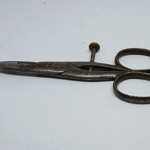 Può includere: Forbici antiche in metallo con una lama appuntita e due fori rotondi per le dita. Le forbici hanno una vite al centro e un aspetto scuro e invecchiato, suggerendo che siano vintage o antiche. Le forbici vengono probabilmente utilizzate per tagliare tessuto o carta.