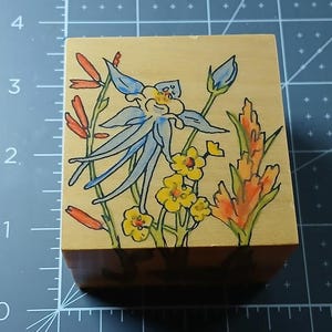 May include: A wooden block with a hand-painted design of blue, yellow, and orange flowers. The flowers are painted in a whimsical style and are surrounded by green leaves.