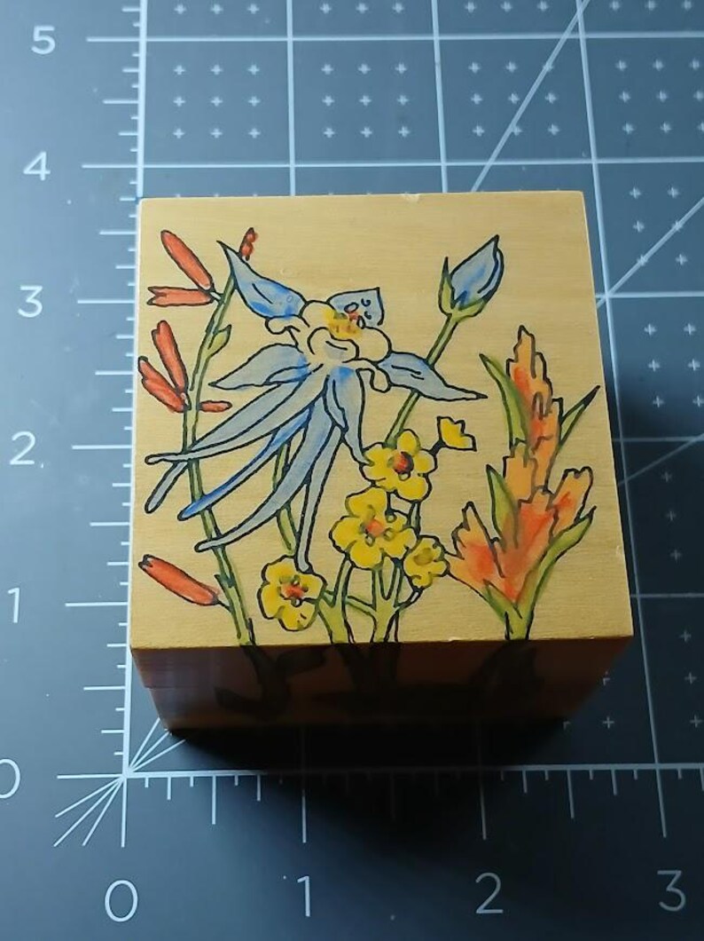 May include: A wooden block with a hand-painted design of blue, yellow, and orange flowers. The flowers are painted in a whimsical style and are surrounded by green leaves.