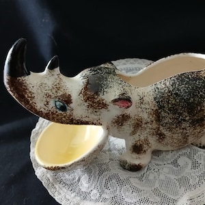 May include: A ceramic rhino planter with a cream, brown, and black speckled finish. The rhino has a yellow interior and a small bowl in front. The planter is placed on a white lace doily, with a dark background.