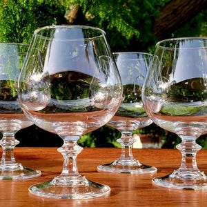 May include: Four clear, stemmed brandy snifters are arranged on a wooden surface. The glasses have a rounded bowl and a short stem. The background features green foliage, creating a contrast with the transparent glassware.