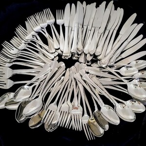 May include: An assortment of shiny silver silverware, including forks, knives, and spoons, arranged in a circular pattern against a black background. The silverware has decorative handles and a polished finish, suggesting a formal dining setting.