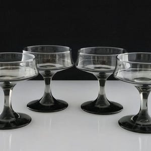 May include: Four clear champagne coupe glasses with a dark gray stem and base. The glasses have a wide, shallow bowl and a rounded rim. The stem is slender, and the base is circular. The glasses are arranged on a white surface.
