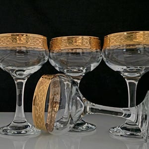May include: Four clear champagne glasses with gold rims. One glass is lying on its side. The glasses have a classic design, with a rounded bowl and a long stem. The gold rims have a decorative pattern.