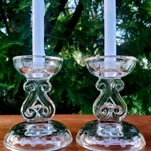 May include: Two clear glass candle holders with white candles. The candle holders have a decorative design on the stem and base, with etched dot patterns. The background is a blurred view of green foliage.