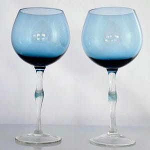 May include: Two elegant wine glasses with a light blue bowl that fades to a darker blue at the base. The stems are clear with a decorative twist. The glasses sit on a clear, round base.