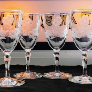 May include: Four clear wine glasses with etched floral designs. The glasses have a tulip shape, with a long stem and a round base. The etched design features a cluster of flowers. The glasses are arranged in a row.