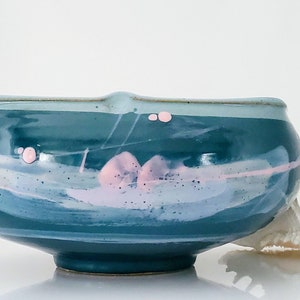 May include: A teal ceramic bowl with a rounded shape and a slightly flared rim. The bowl features abstract pink and white painted designs, with a speckled texture. The exterior is a glossy finish, and the bowl sits on a white surface.