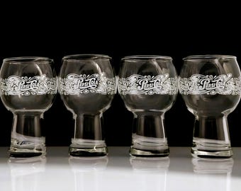 May include: Four clear glass Pepsi Cola branded beer glasses. Each glass has a bulbous bowl and a short stem. The Pepsi Cola logo is printed in white around the top of each glass. The glasses are arranged in a row against a black background.