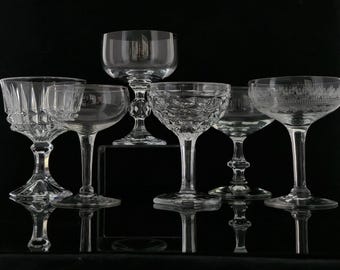 May include: An assortment of clear glass champagne glasses with various designs. The glasses have different stem and bowl shapes, including etched and cut patterns. The glasses are arranged against a black background.
