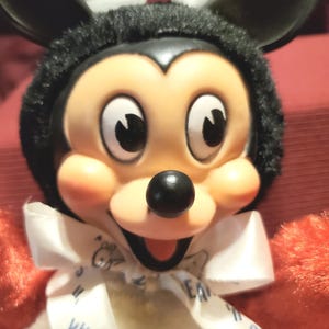 May include: A plush Mickey Mouse doll with a black head and ears, a tan face, and a black nose. The doll has a white bow tie with the word "Tweak" visible. The doll's arms are red.