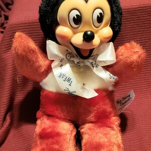 May include: A vintage Mickey Mouse plush toy with a black head and ears, a red body, and white accents. The toy has a white ribbon around its neck with the word "Tweak" printed on it. A "Walt Disney Character" tag is attached.
