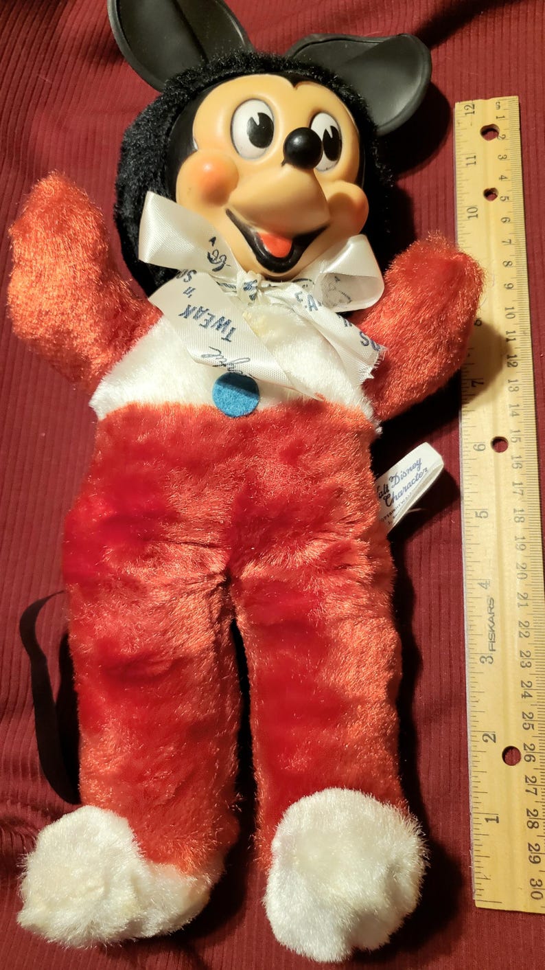 May include: Vintage Mickey Mouse plush doll with a black head, orange cheeks, and red plush body. The doll has a white collar with the text "Tweak" and a blue button. A tag reads "Walt Disney Character".